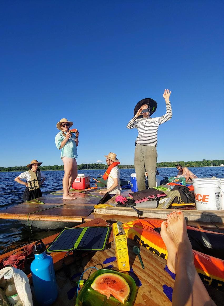 a close up of a similar scene from the previous photo with a man in a black hat and blue striped shirt waving and a woman in a teal shirt, hat and sunglasses eating watermelon